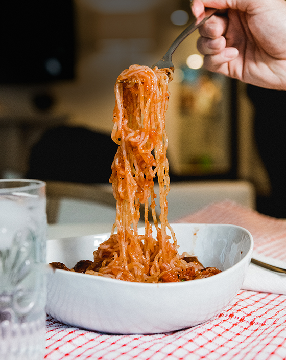 A fork lifting spaghetti with sauce from a white bowl on a table.