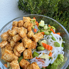 Bowl of tofu, noodles, and mixed vegetables with greenery in the background.