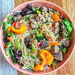 Bowl of noodles with vegetables and meat chunks on a wooden table.