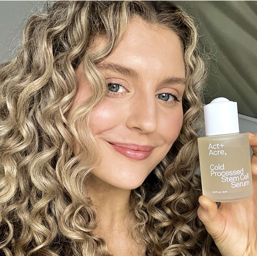 Person with curly hair holding a bottle of Act+Acre Cold Processed Stem Cell Serum.