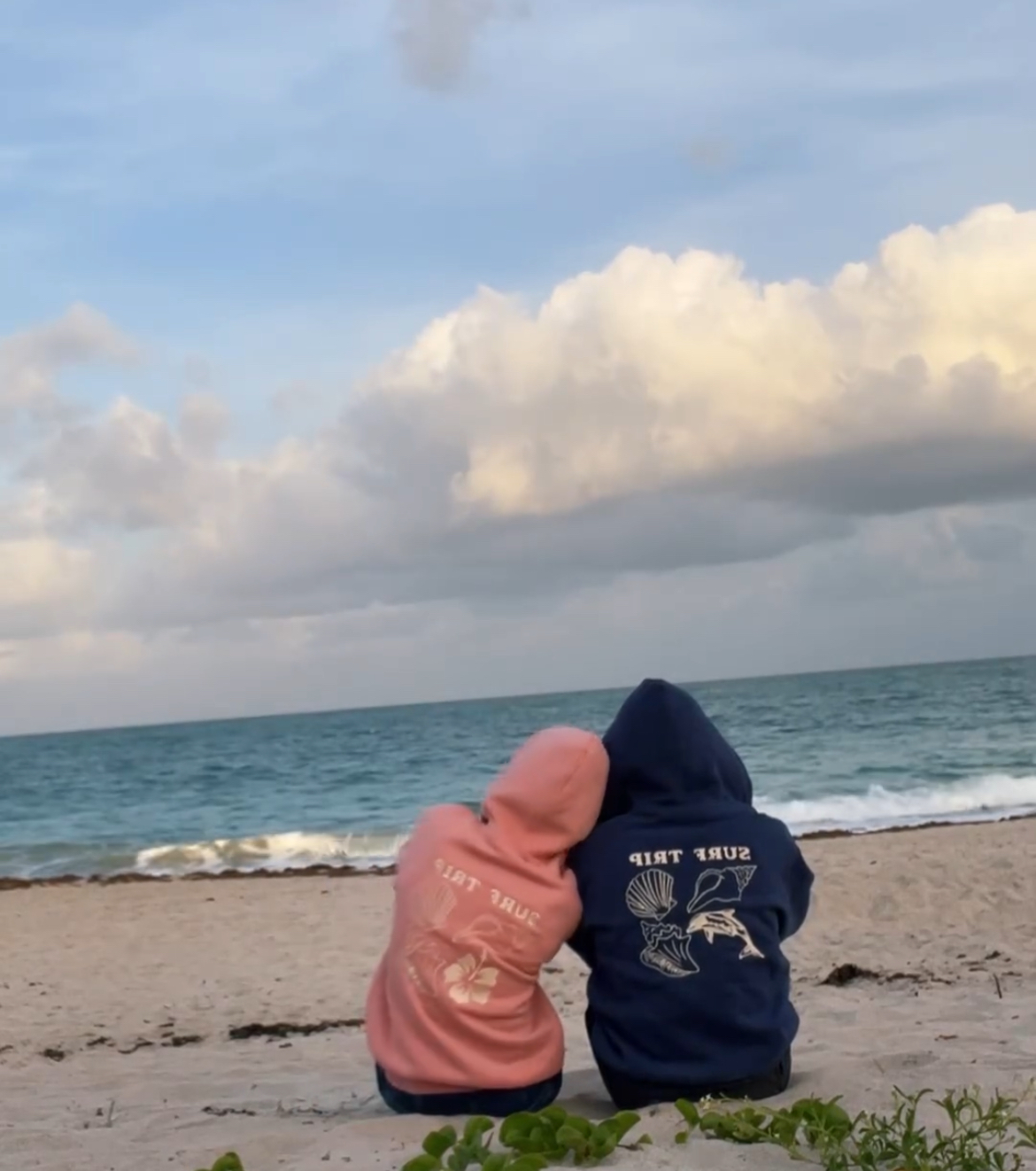 Two people in hoodies sitting on a beach, facing the ocean.