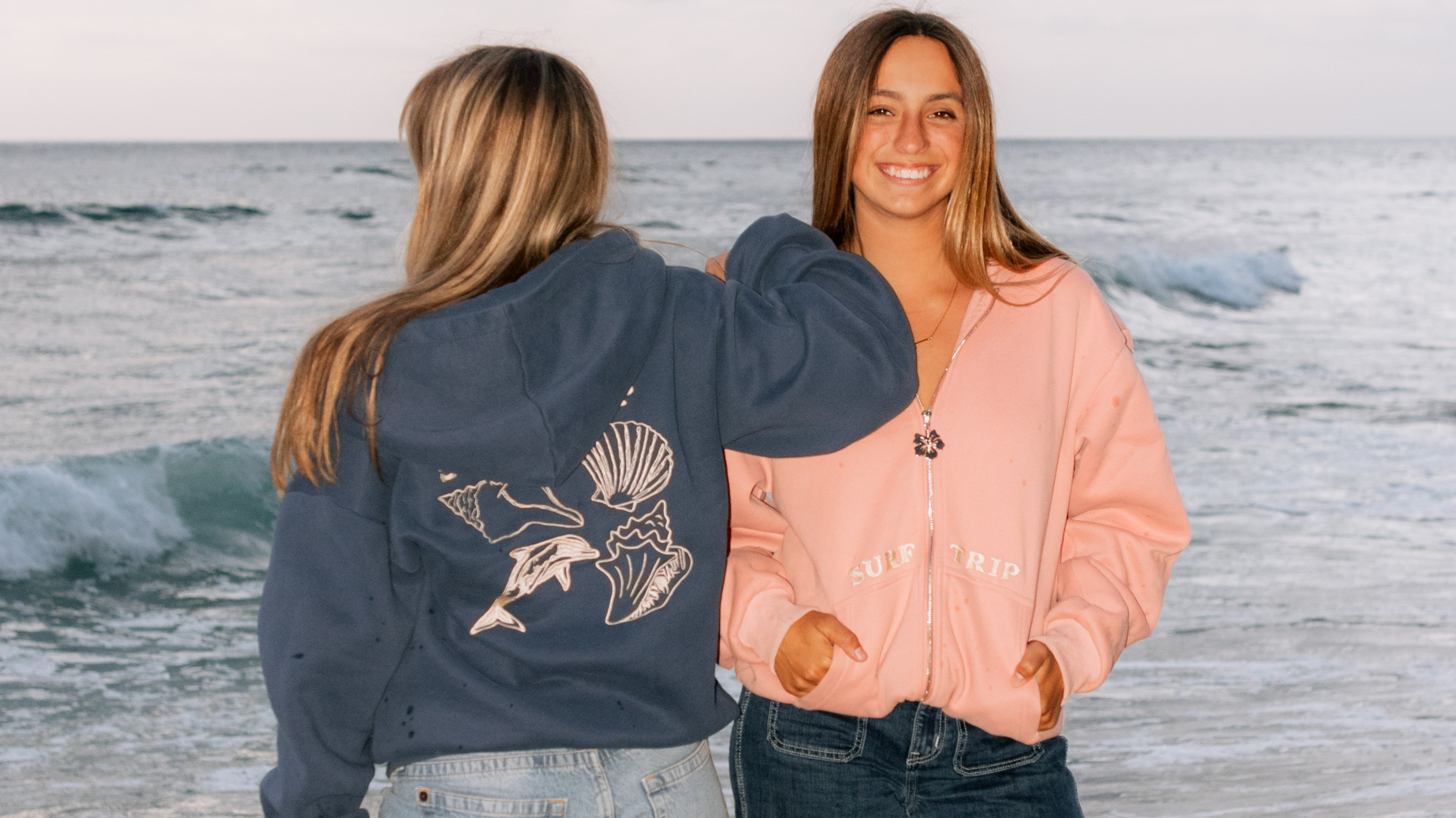 Two people in hoodies standing by the ocean, one facing forward, the other backward.
