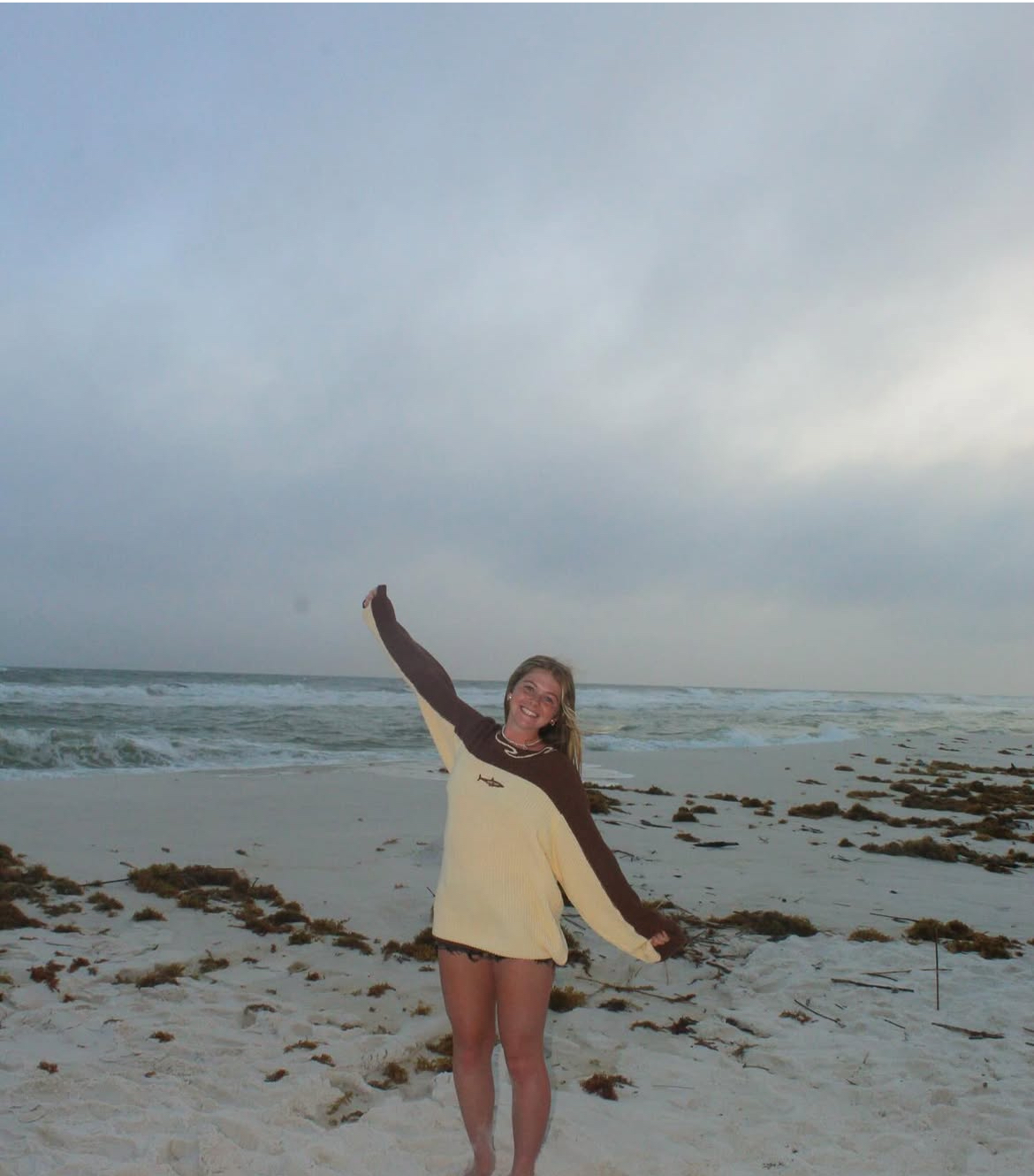 Person smiling on a beach with seaweed and cloudy sky.