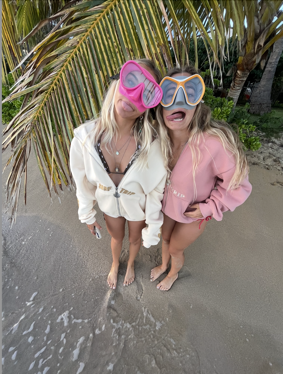 Two people on a beach wearing snorkel masks and hoodies, making funny faces.