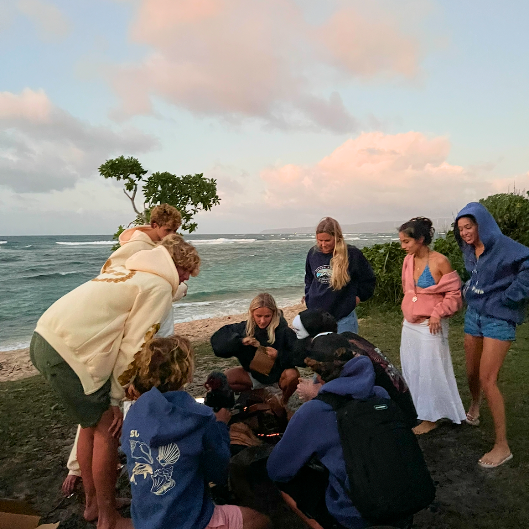 Group of people gathered around a small fire on a beach at sunset.