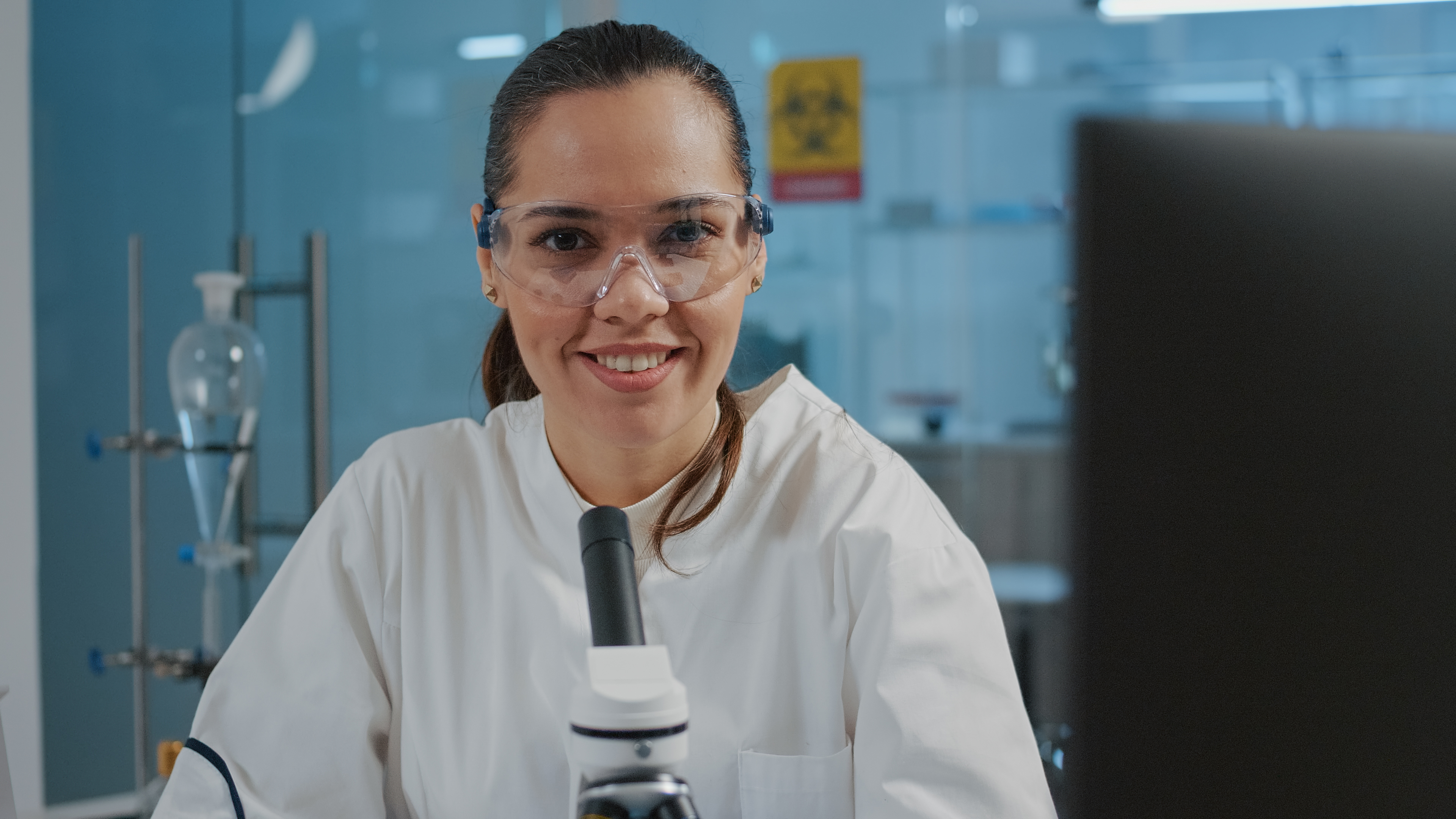 Smiling person in a lab coat and goggles with a microscope.