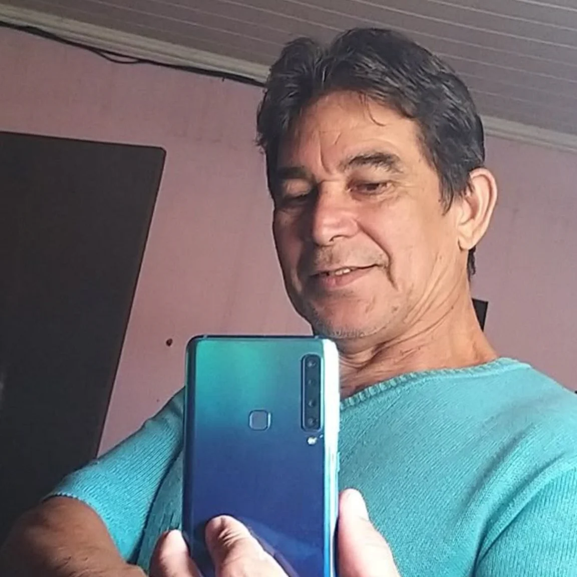 A middle-aged man in a turquoise sweater holds a blue smartphone, appearing to take a selfie.