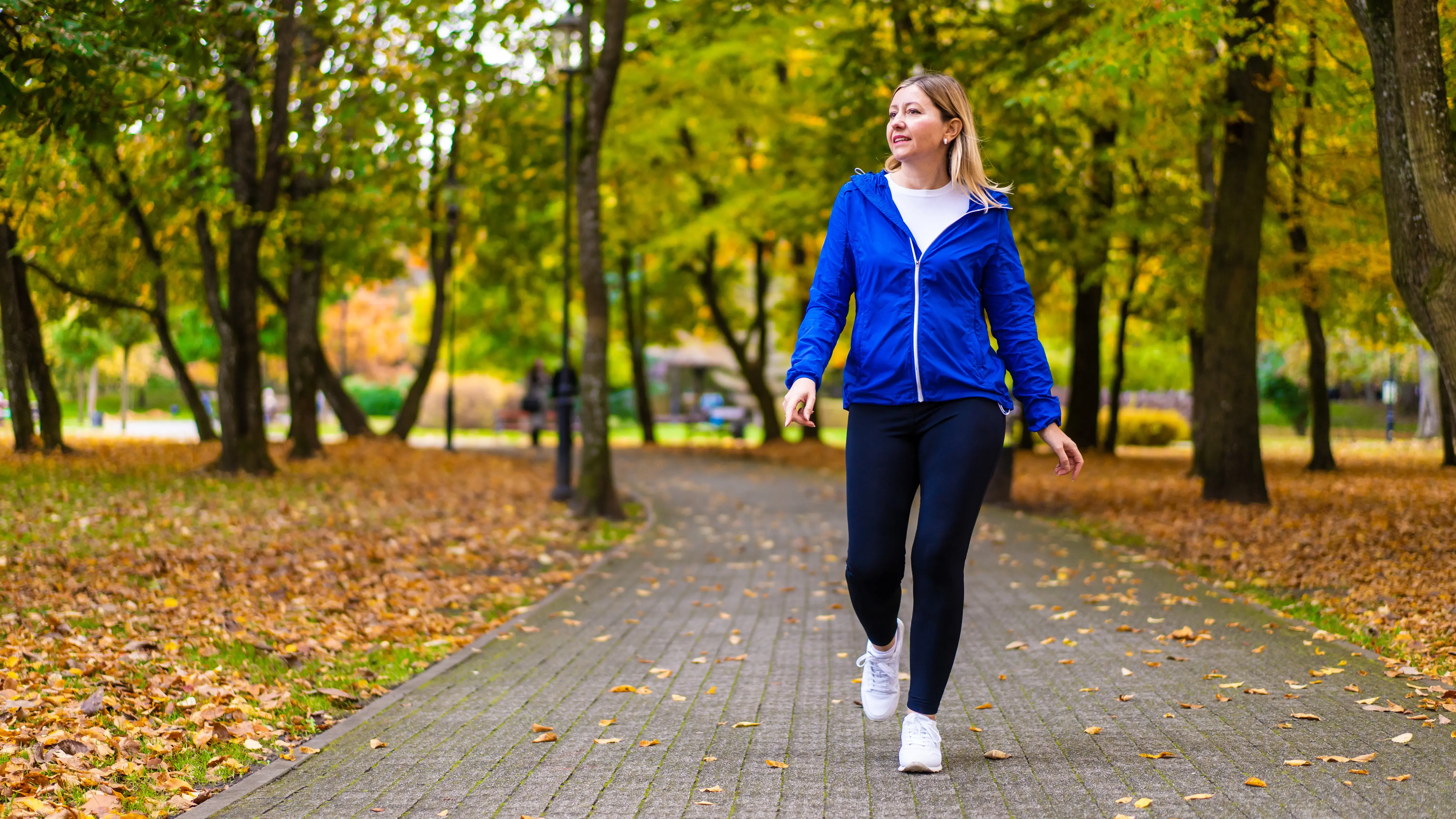 A woman in a blue jacket walks on a paved path through a park in autumn.
