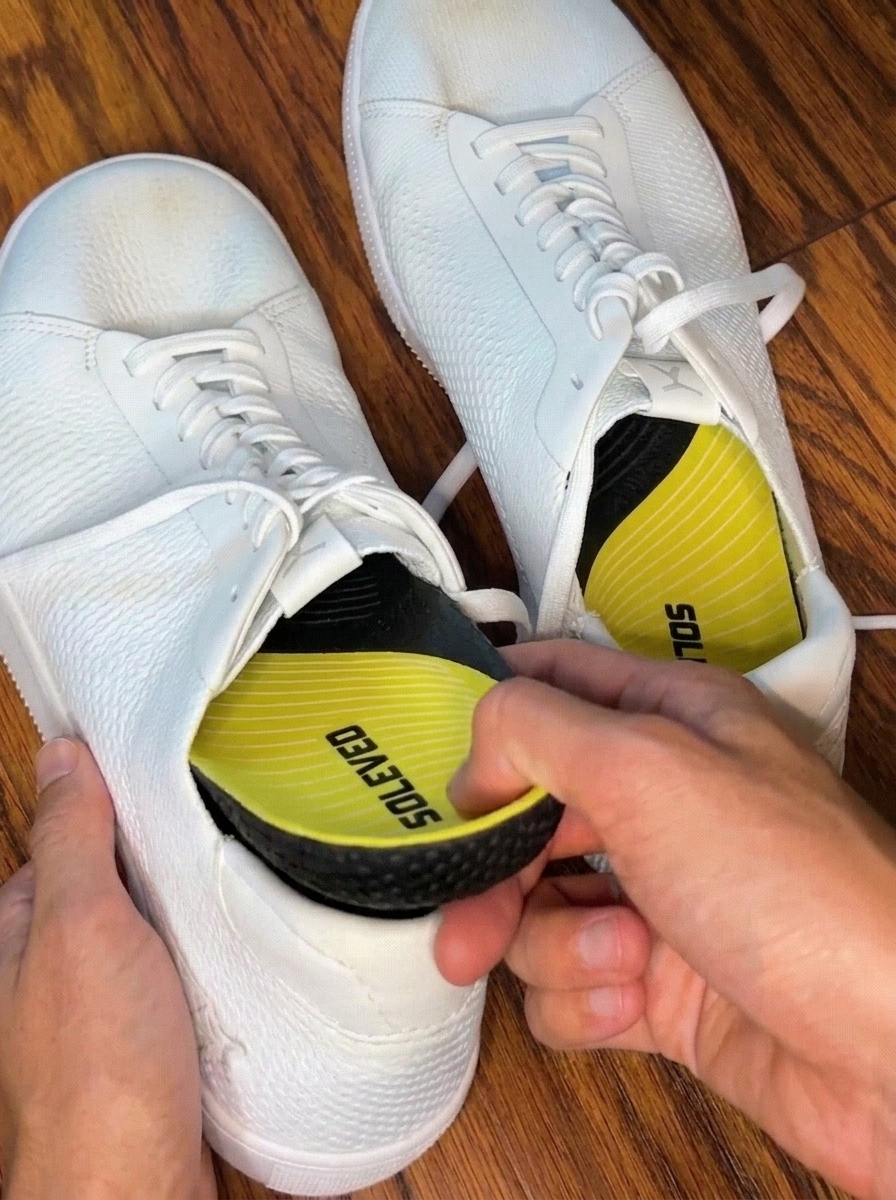 A person's hands inserting a yellow and black SOLEVED insole into a white textured sneaker.