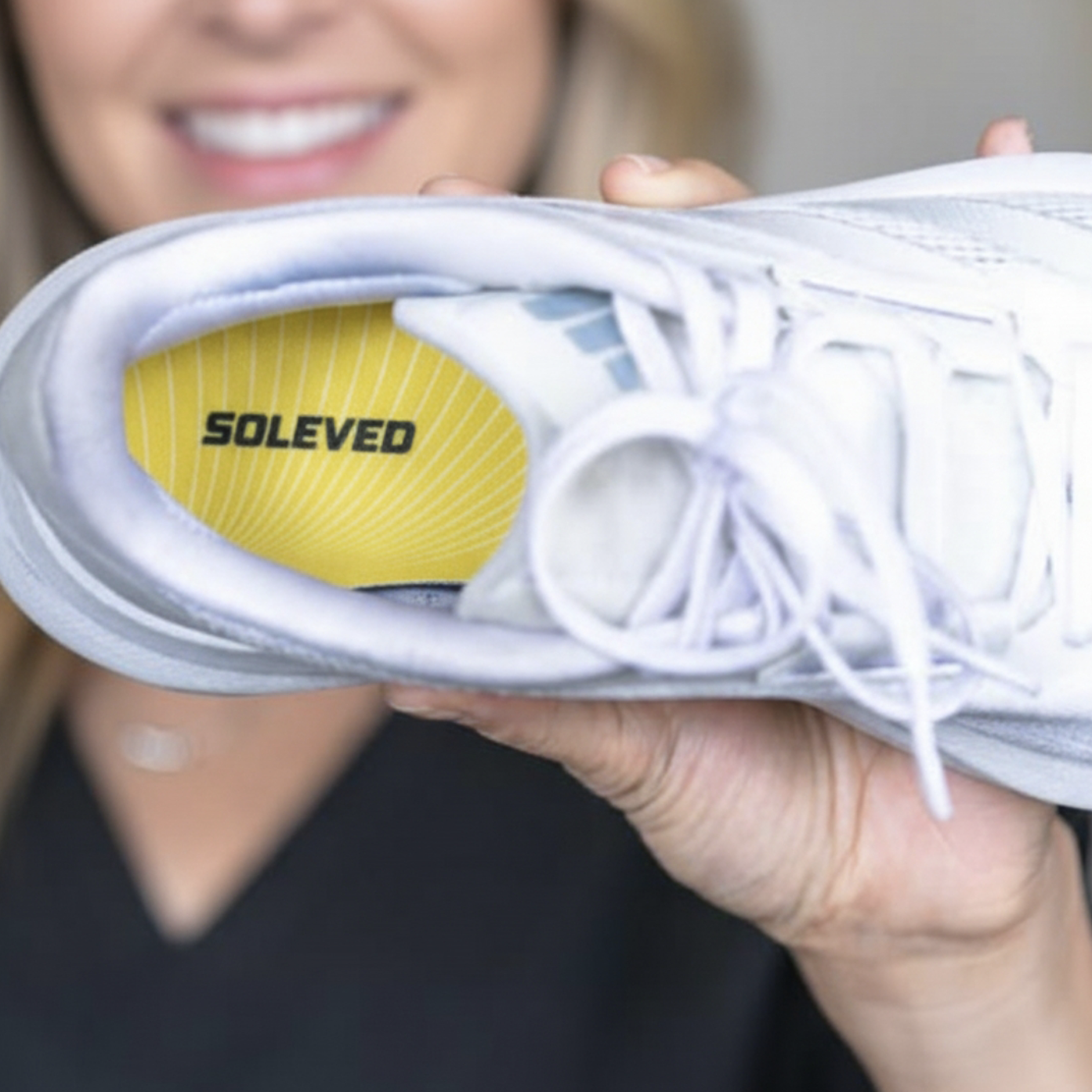 A person holds up a white sneaker, showing the yellow SOLEVED insole inside.