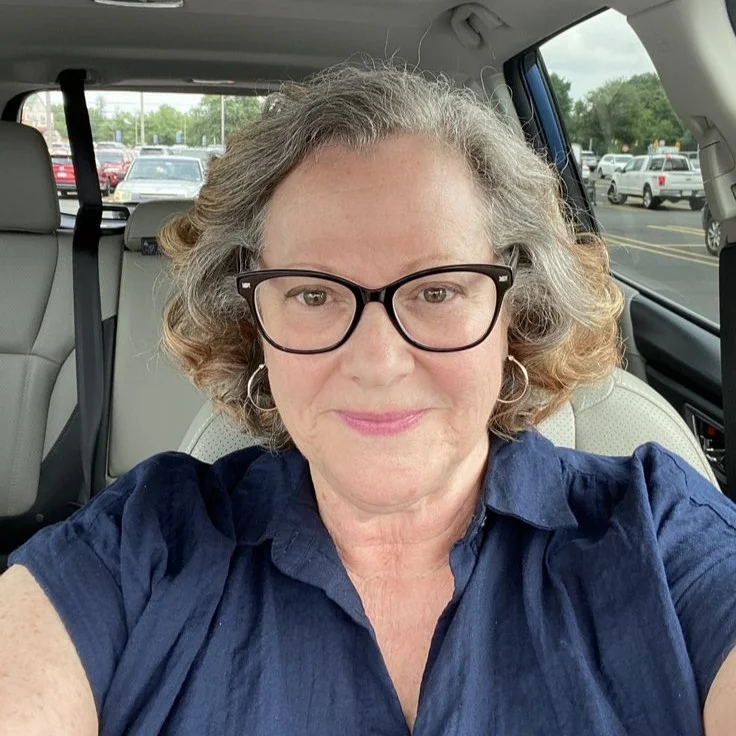 A woman with graying curly hair and glasses smiles while sitting in the driver's seat of a car.