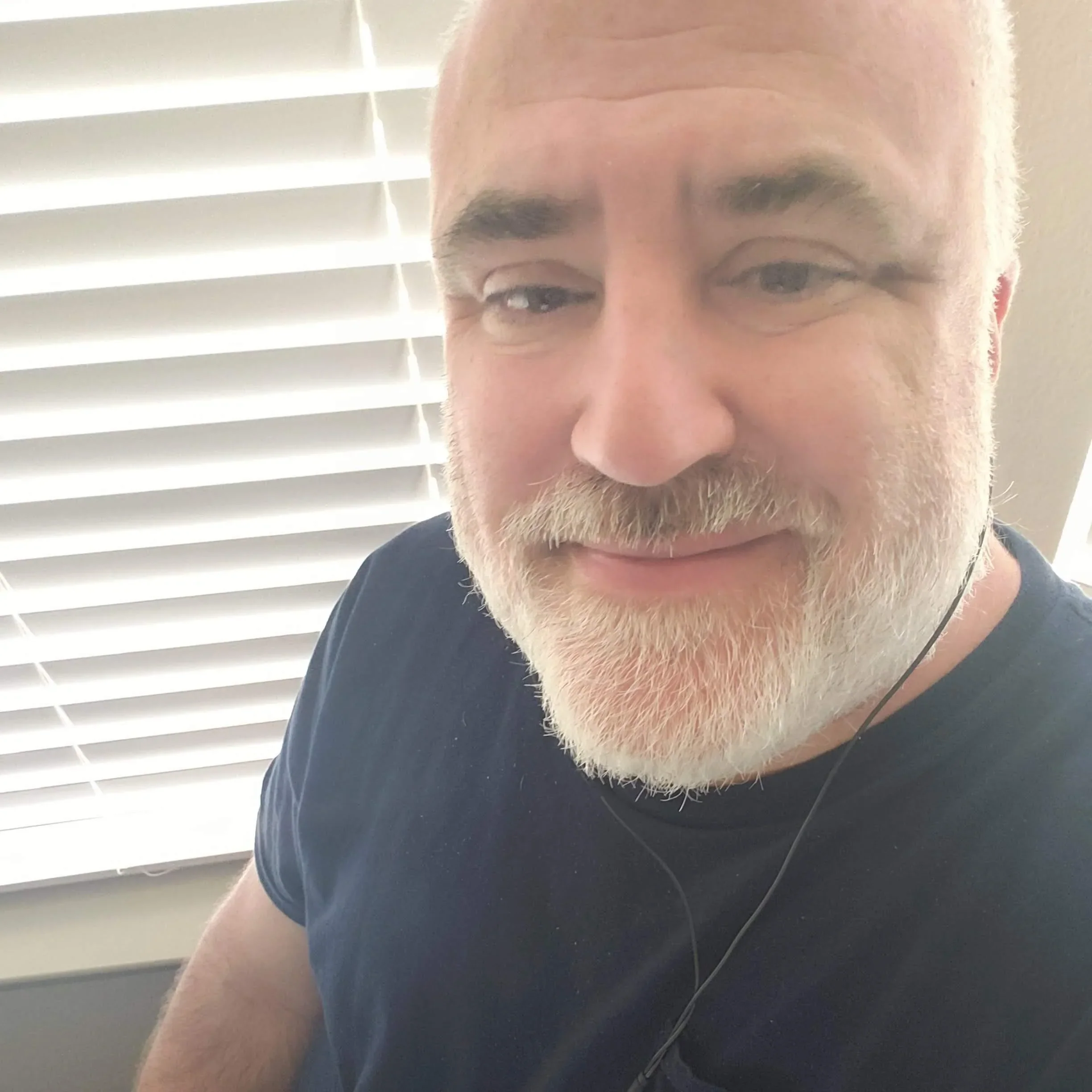 A close-up of a smiling man with a white beard, wearing a dark blue t-shirt.