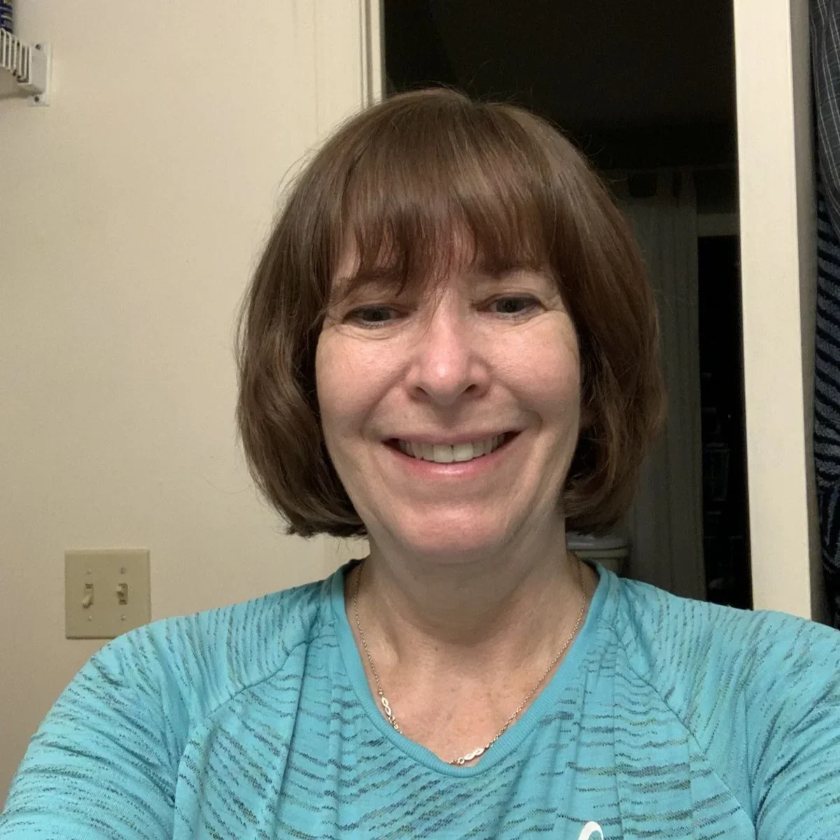 A smiling woman with short brown hair and bangs, wearing a blue patterned shirt, takes a selfie indoors.