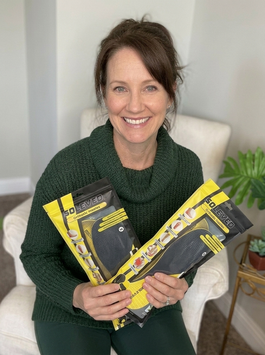A smiling woman in a green sweater holds two packages of orthotic insoles while sitting in a chair.