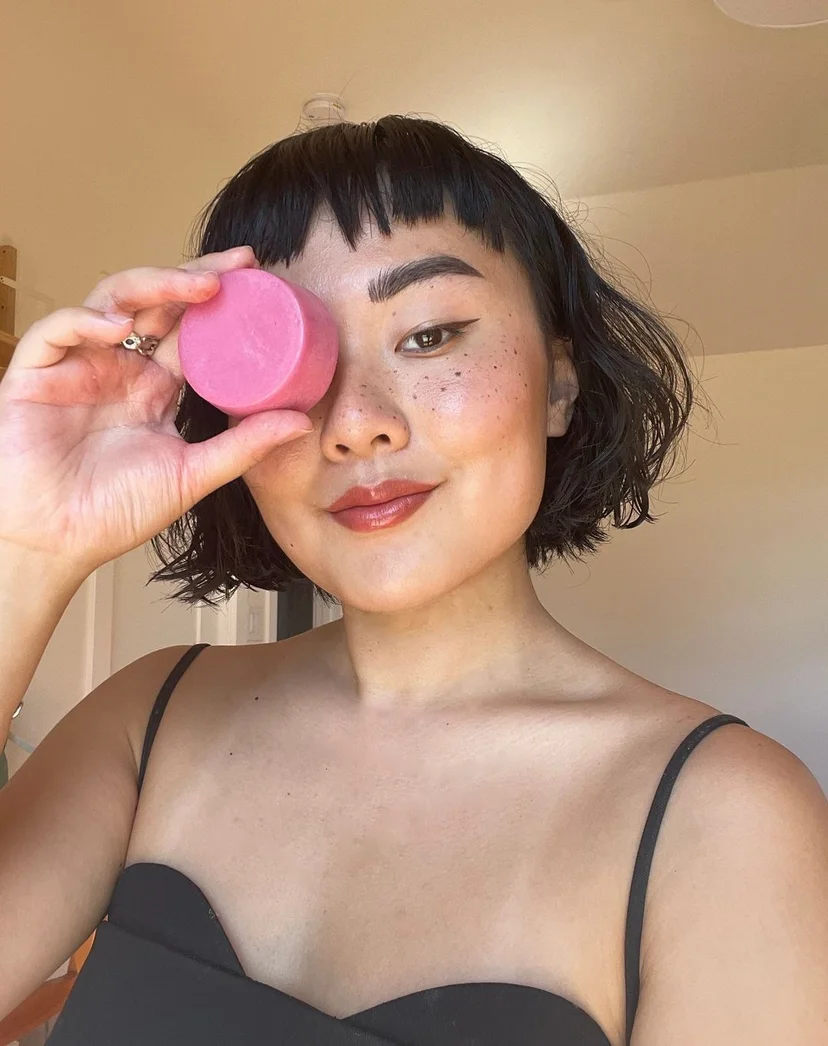 Person holding a pink round object near their eye, smiling.