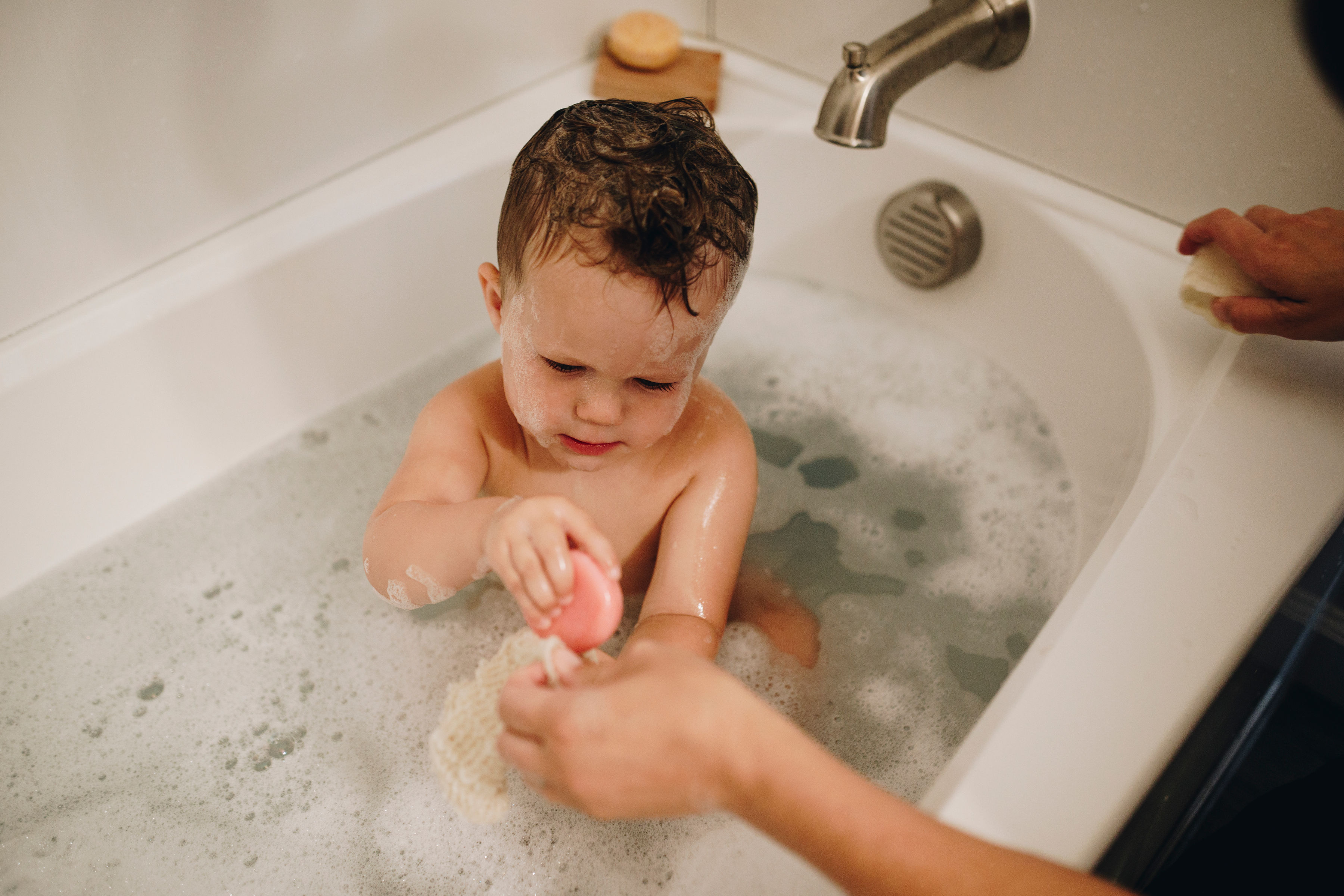 Child in a bathtub with bubbles, holding a pink toy.