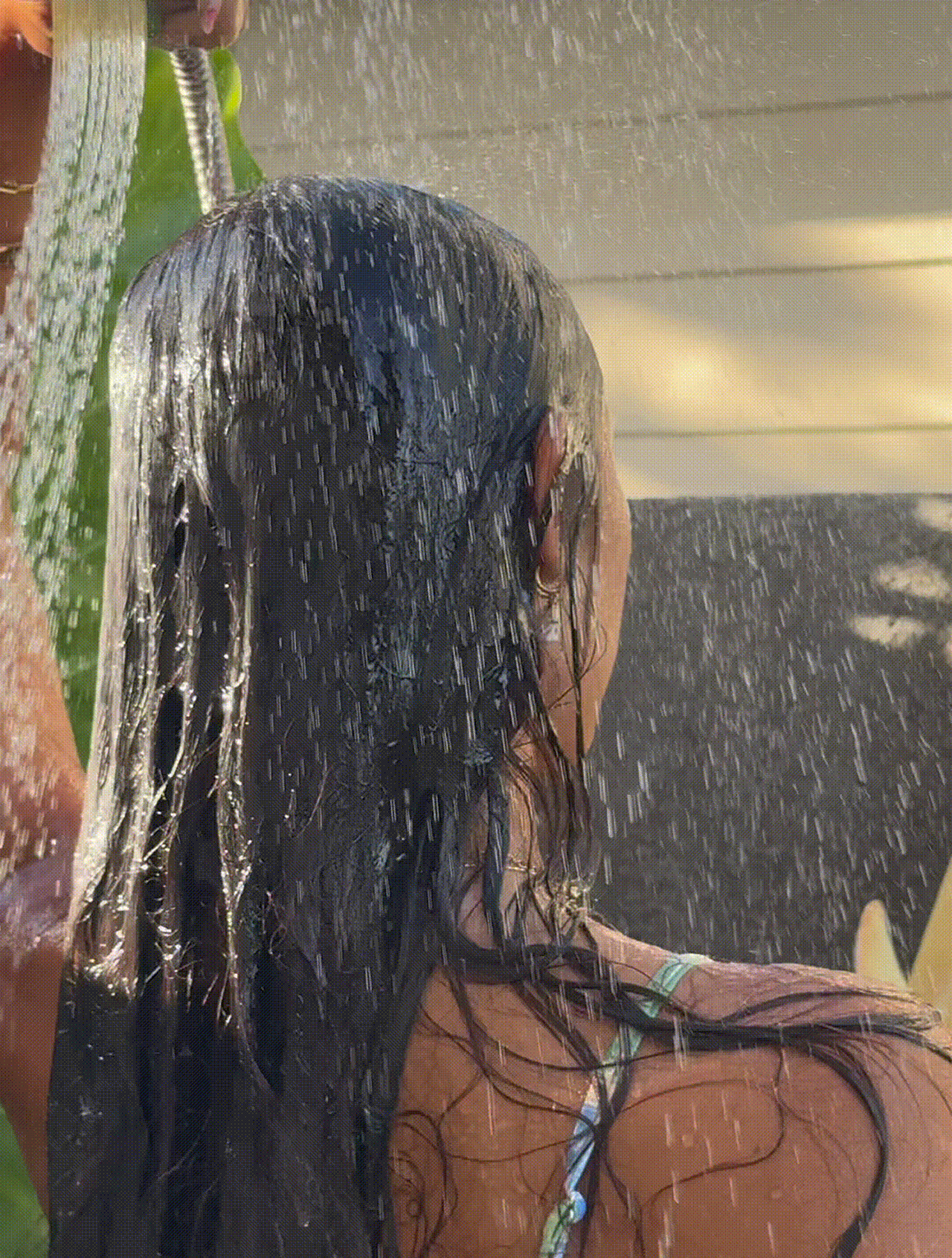 A person with long, dark wet hair standing under an outdoor shower, seen from behind.