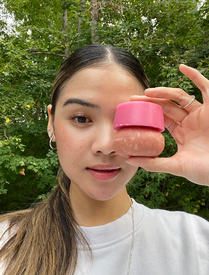 Person holding two stacked round objects, one pink, in front of their face outdoors.