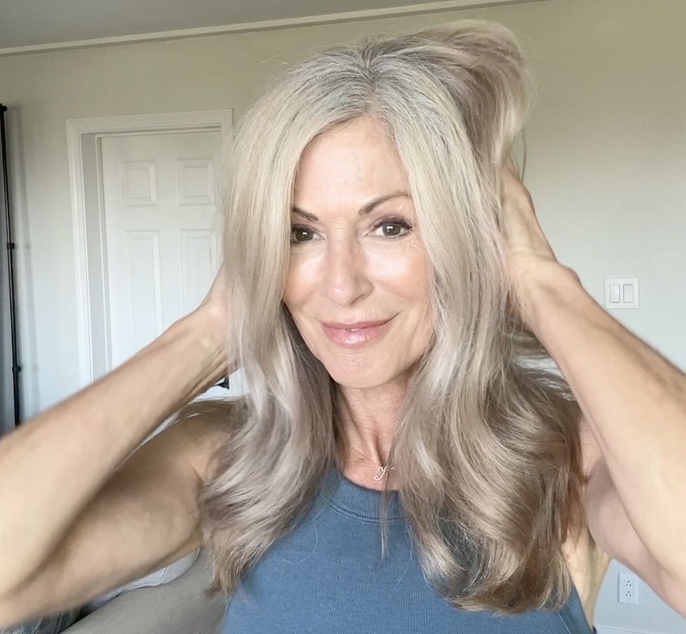Person with long, light hair touching their head, smiling in a room with neutral colors.