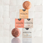 A stack of four shampoo and conditioner bar boxes with two unwrapped bars on a reflective surface.