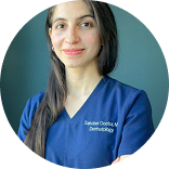 Person in blue medical scrubs with long hair against a neutral background.