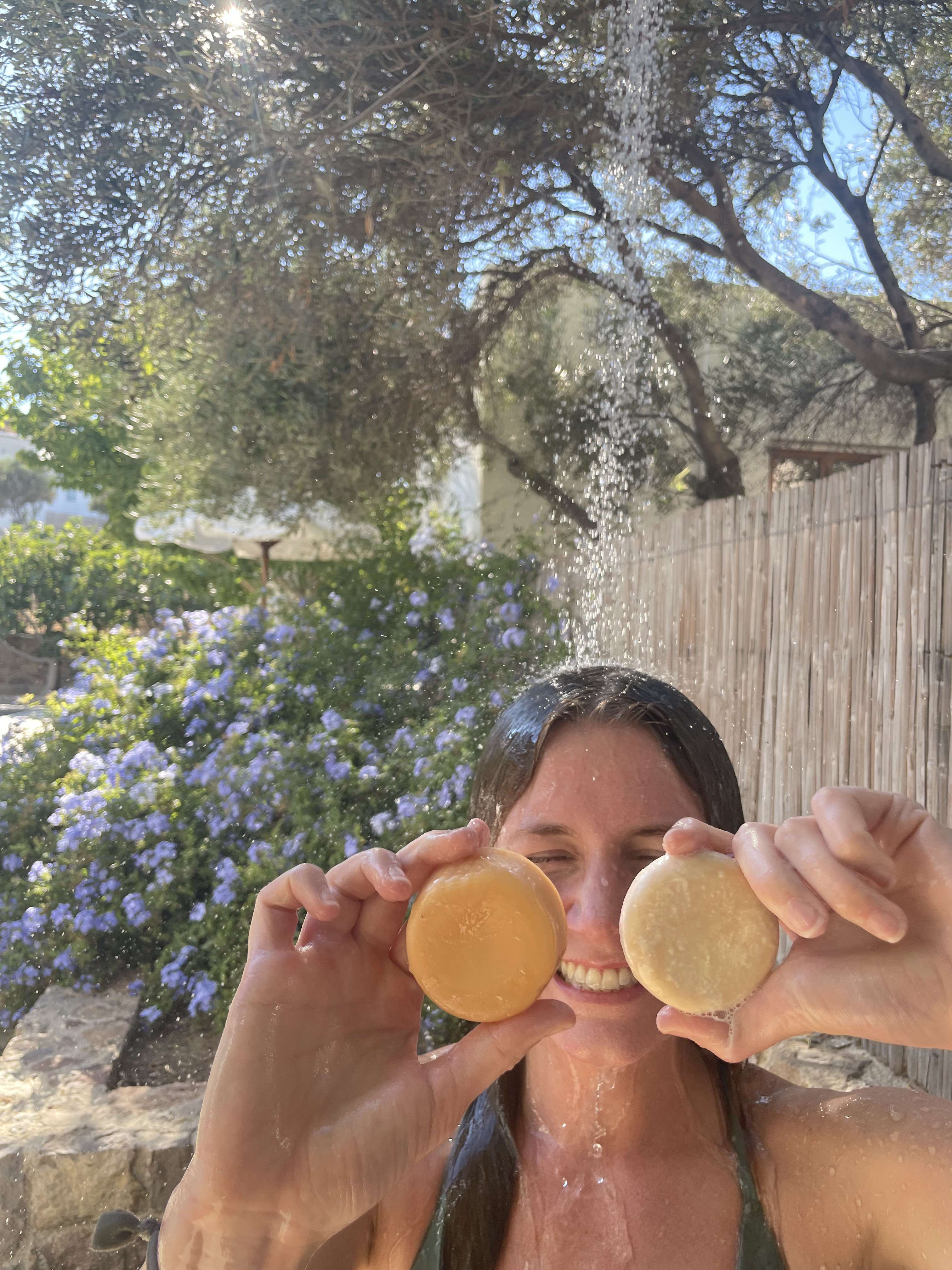 A smiling woman in an outdoor shower holds two round soap bars in front of her eyes.