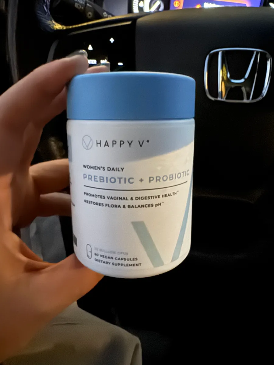 Hand holding a jar of Happy V prebiotic and probiotic supplements inside a car.