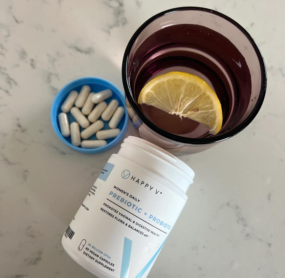 Container of supplements next to water with lemon and a dish of capsules on a marble surface.