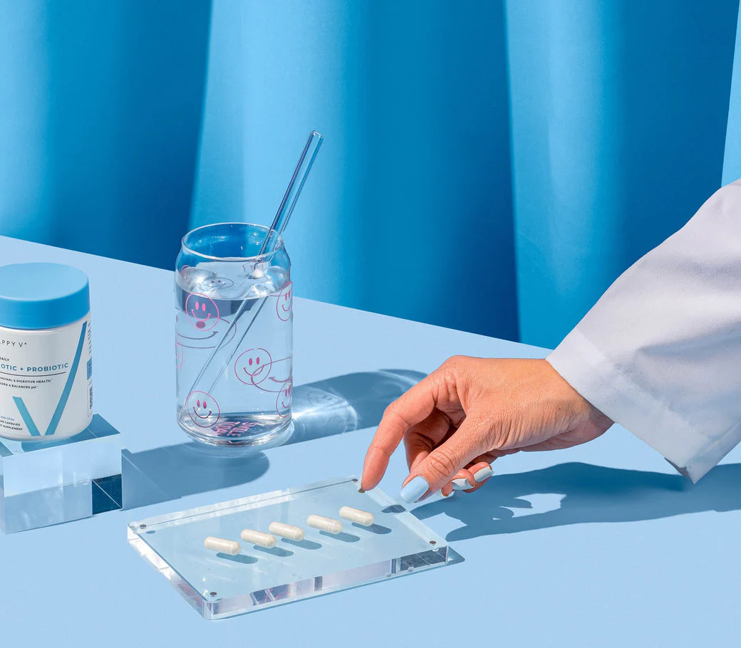 A hand taking a capsule, next to a glass of water and a probiotic supplement container on a blue surface.