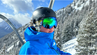 Person in blue jacket and helmet on ski lift with snowy mountains.