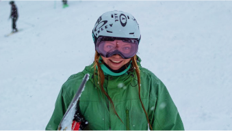 Person in green jacket and helmet holding skis on a snowy slope.