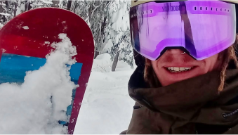 Person in snow gear holding a snowboard with snow-covered trees in the background.