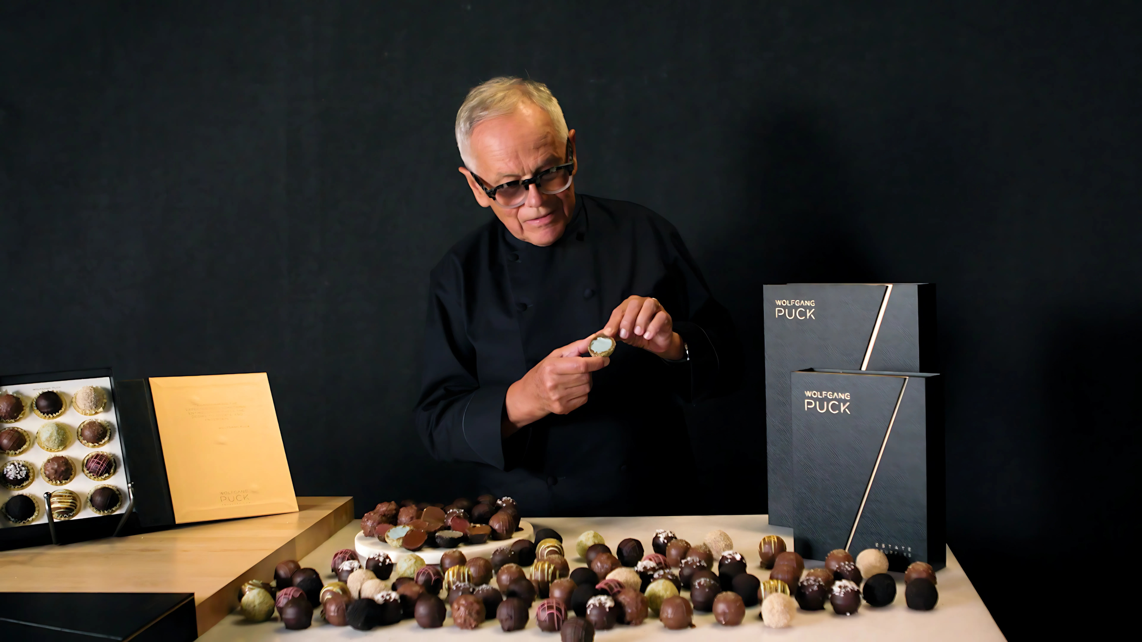 Person in black clothing with chocolates on a table, black boxes labeled 'Wolfgang Puck'.