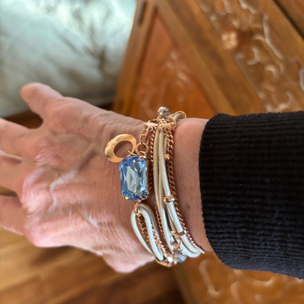 A wrist wearing a multi-strand bracelet with white cords, rose gold chains, and a blue gemstone charm.