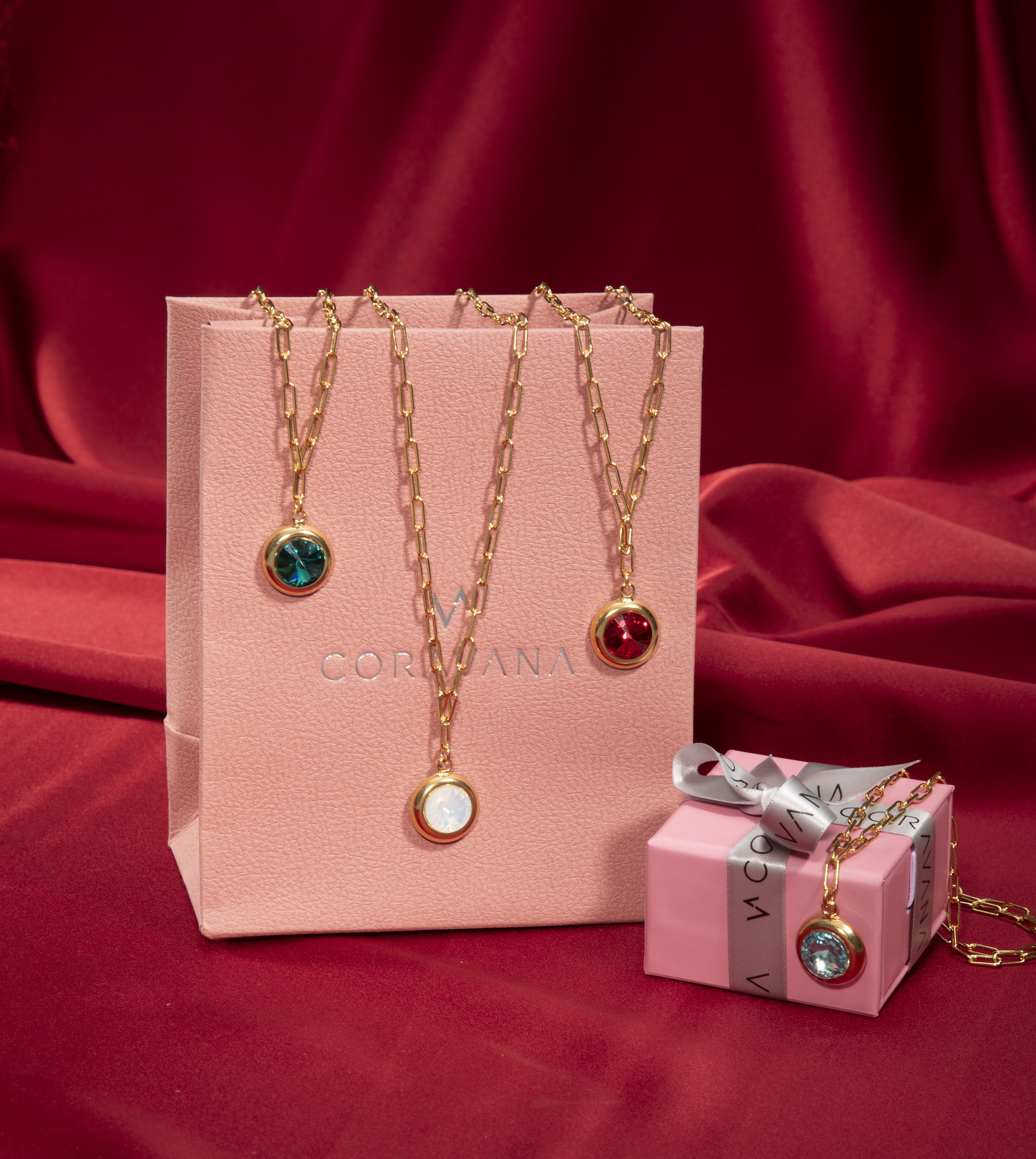 Three gold chain necklaces with colorful pendants on pink bags against a red fabric background.