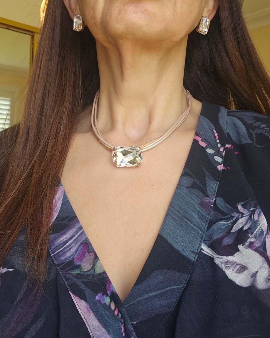 A woman with long brown hair wearing a crystal necklace and matching earrings with a floral top.
