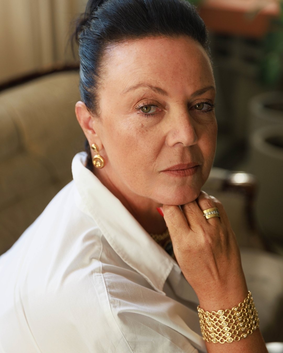A woman with dark hair in a white shirt and gold jewelry rests her chin on her hand.
