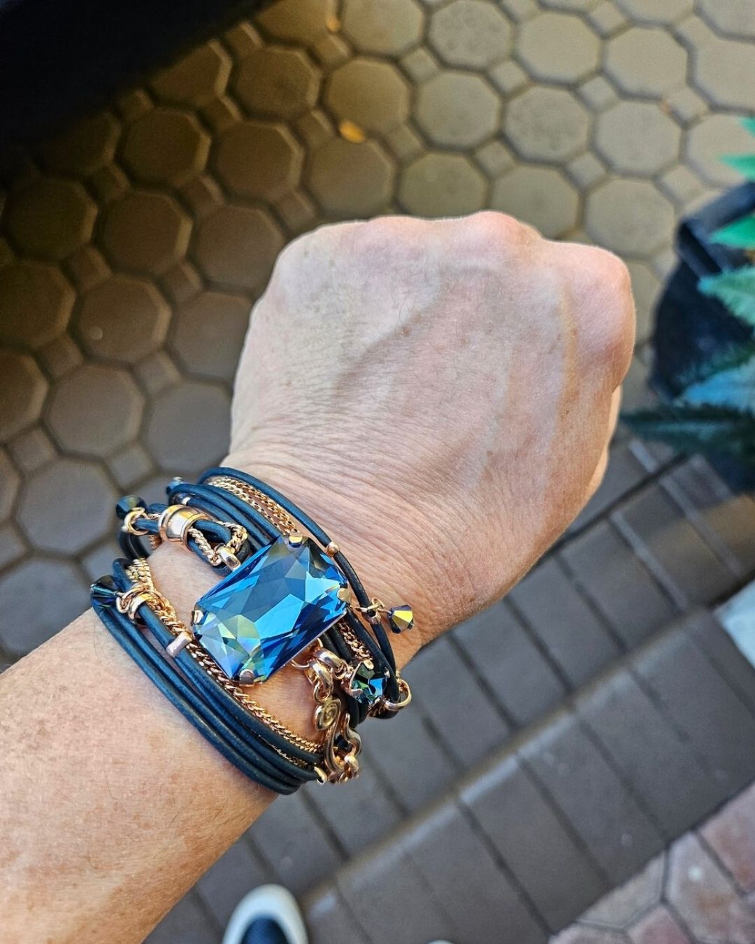 A person's wrist wearing a multi-strand bracelet with a large, rectangular, faceted blue stone and gold-colored chains.