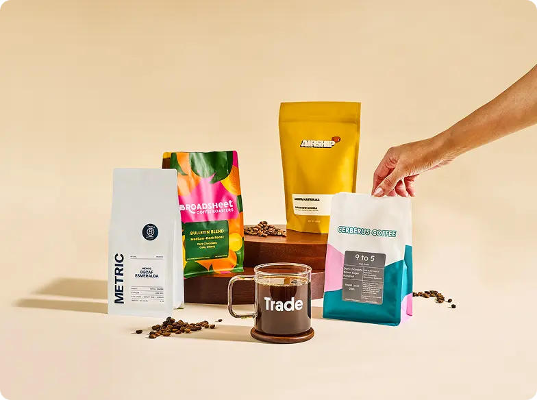 Various coffee bags displayed around a glass mug filled with black coffee. The mug reads 'Trade' and sits on a wooden coaster. An arm reaches out to grab one of the bags from out of frame.
