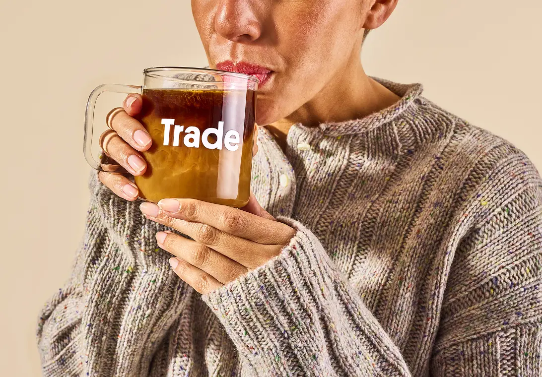Closeup of a woman sipping coffee from a glass mug with 'Trade' on the front.
