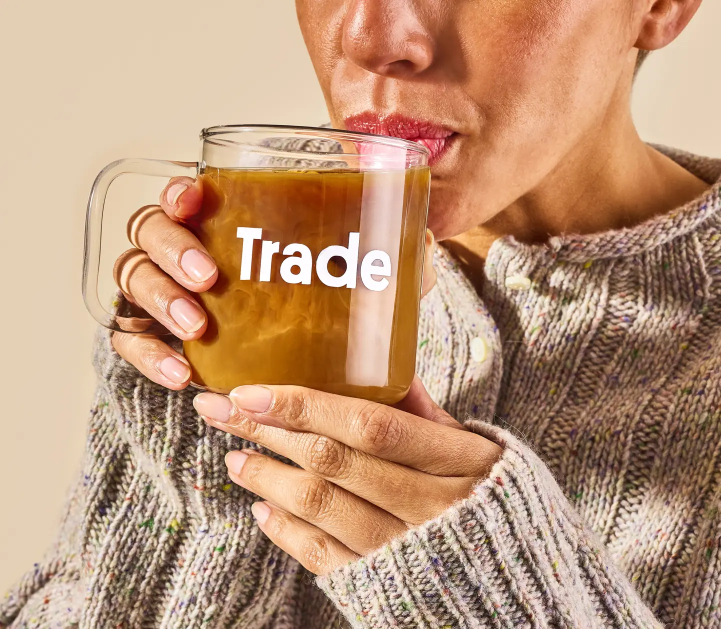 Closeup image of a woman sipping coffee from a glass mug with 'Trade' on the front.