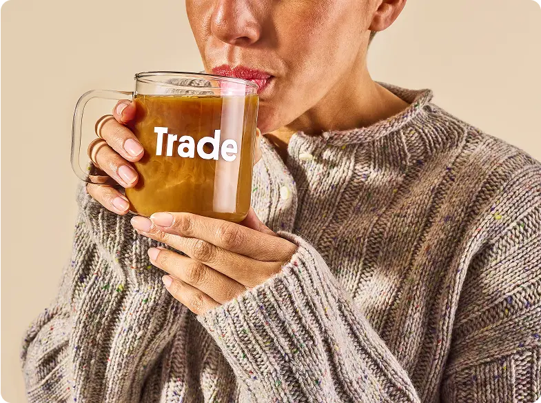 Closeup of a woman in a knit sweater sipping from a glass mug filled with coffee. The mug reads 'Trade'.