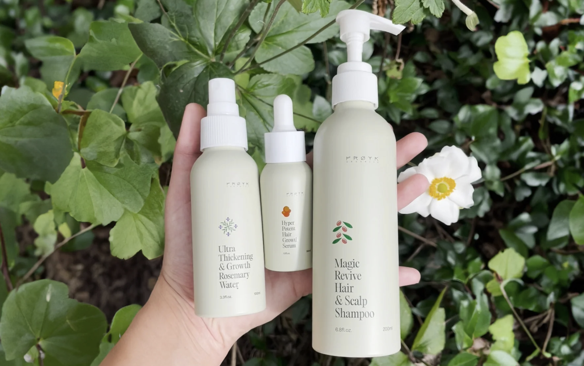 Hand holding three hair products with green leaves and white flower background.