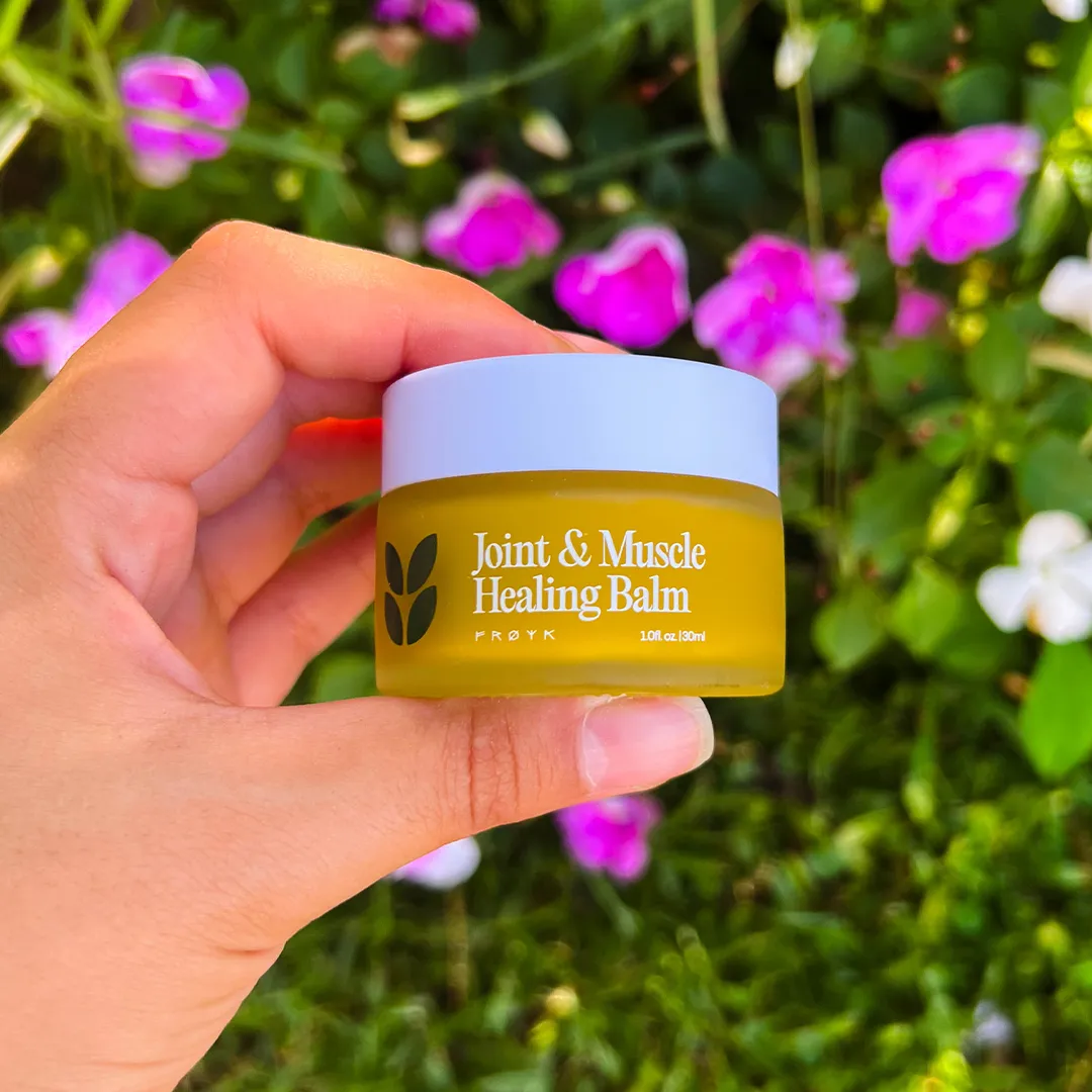 Hand holding a jar of Joint & Muscle Healing Balm with flowers in the background.