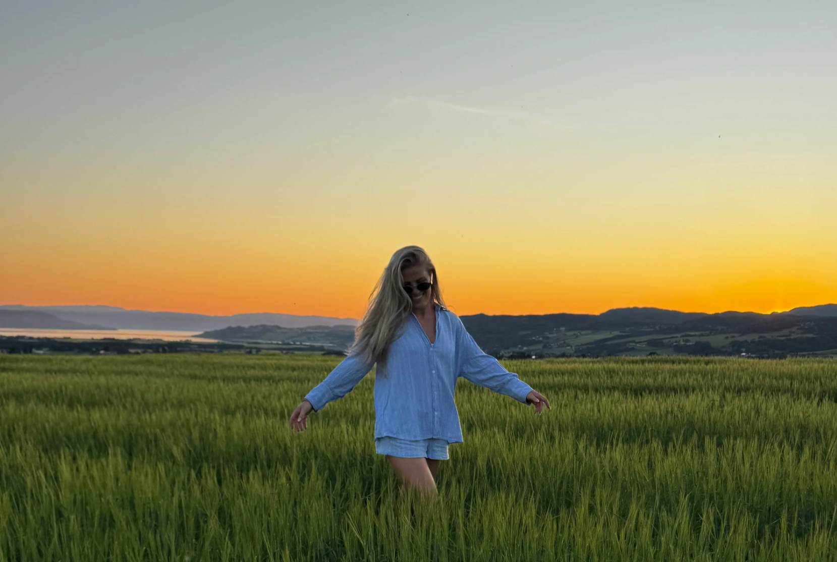 Person in a field at sunset, wearing a blue shirt and sunglasses.