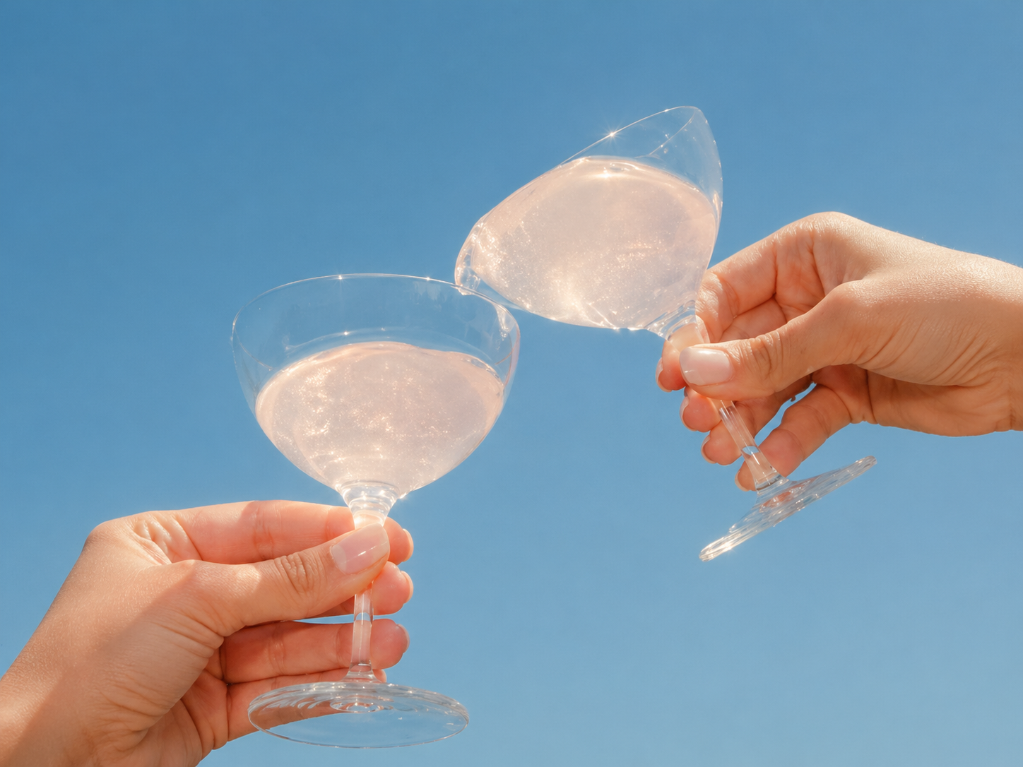 Two hands clinking coupe glasses with a shimmery pink drink against a clear blue sky.