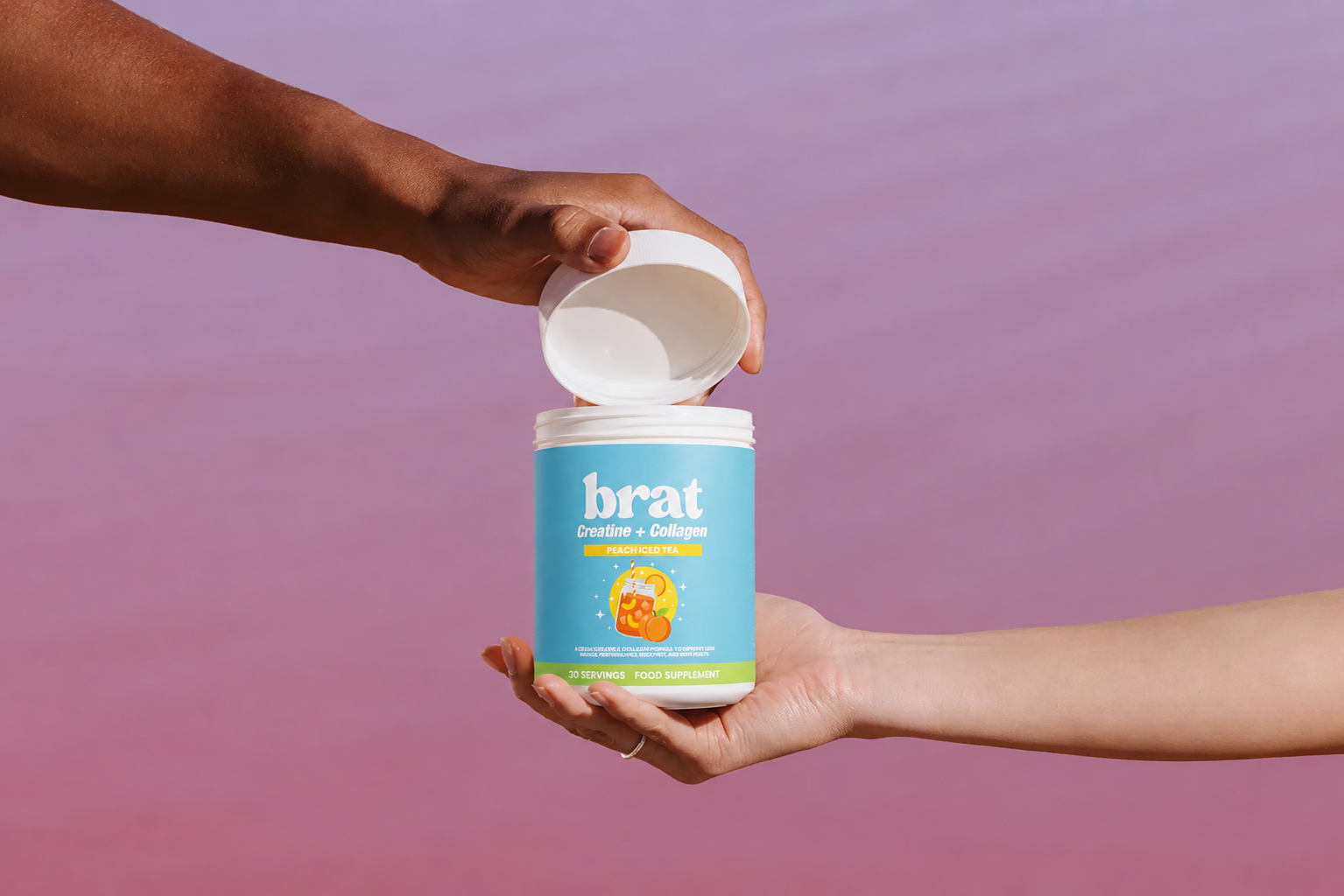 Two hands opening a jar of Brat Creatine + Collagen peach iced tea supplement against a pink background.