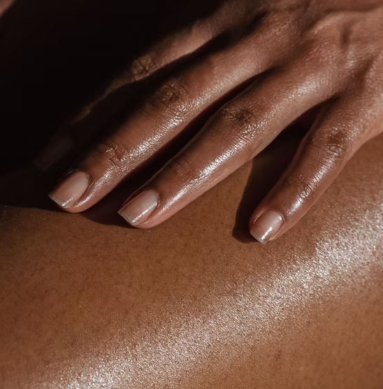 A close-up of a manicured hand resting on glowing, oiled skin under dramatic lighting.