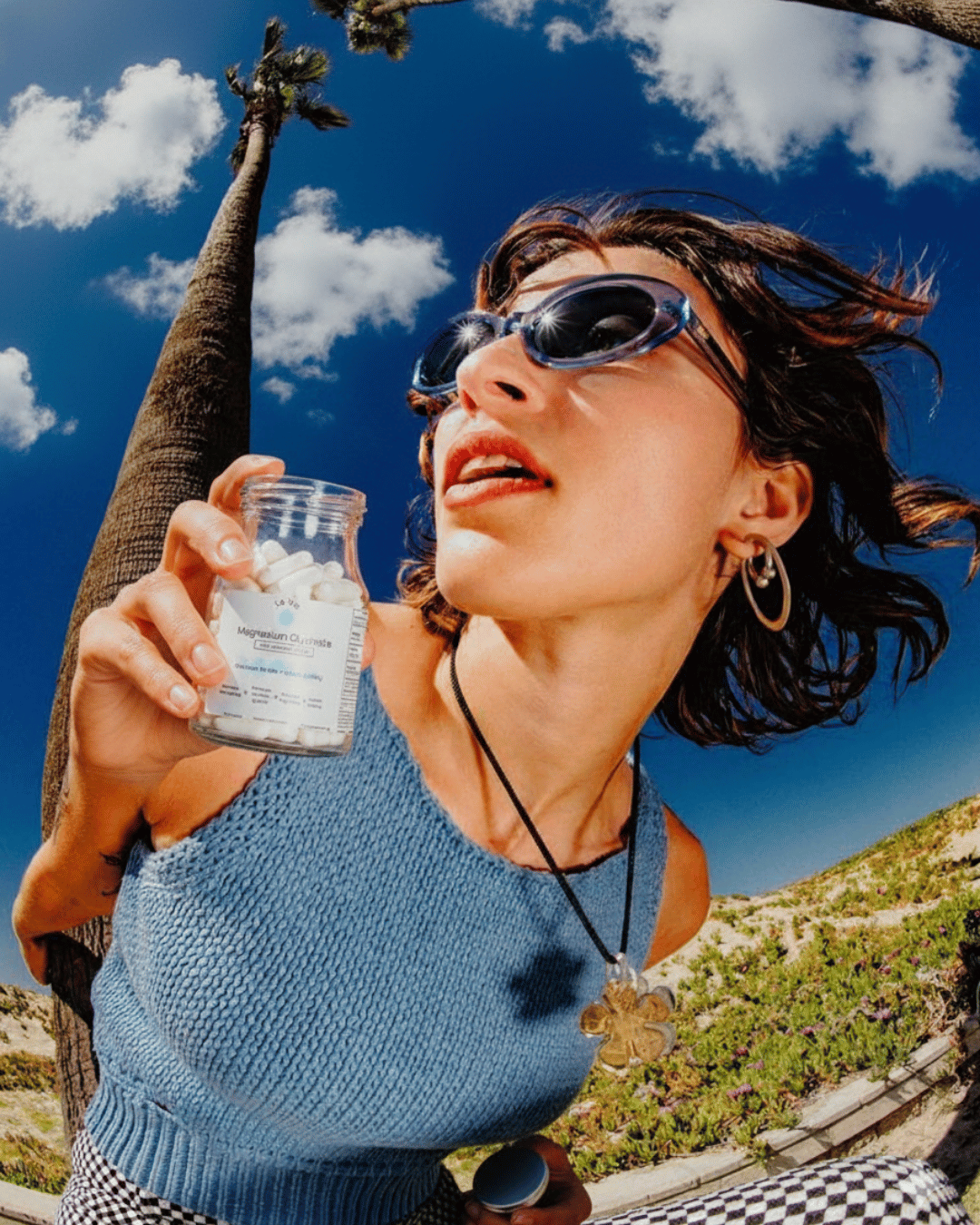 Person holding a supplement bottle near a palm tree under a blue sky.