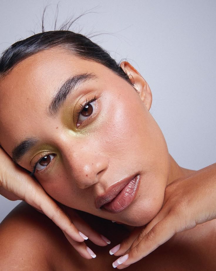 Close-up of a person with green eyeshadow and glossy lips, resting head on hands.